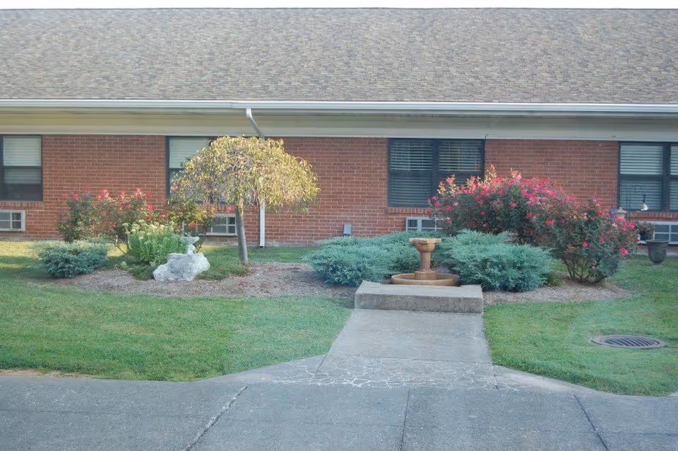 Front lawn and walkway of a brick senior living building with shrubs, flowering bushes, a small fountain, and a small tree.