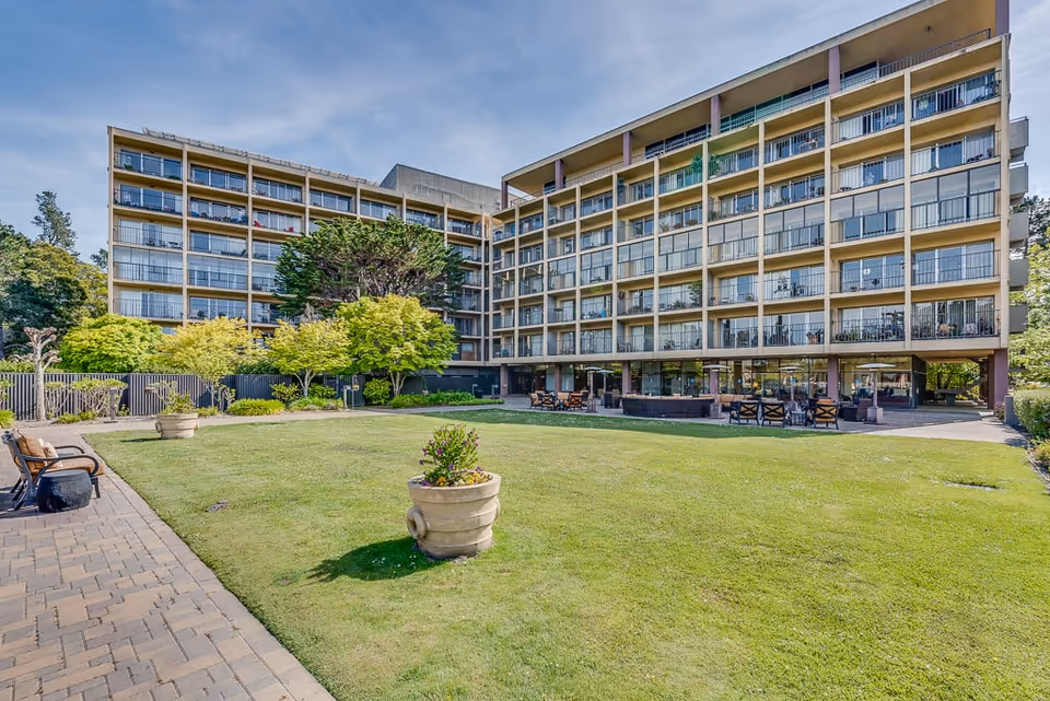 Exterior view of a multi-story senior living facility building with balconies overlooking a well-maintained grassy courtyard. The courtyard features potted plants, benches, and outdoor seating areas under a clear blue sky.