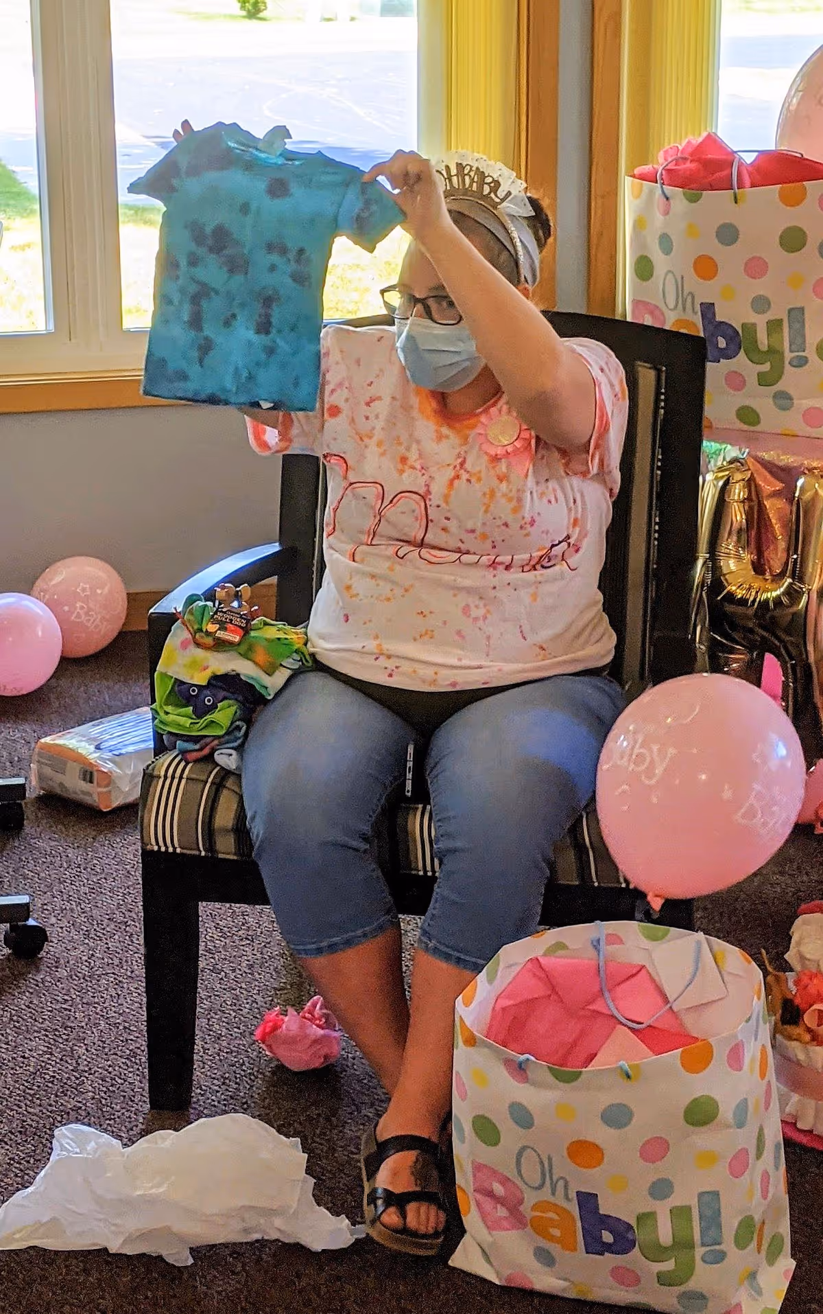 A woman wearing a birthday tiara and a face mask is sitting on a chair indoors, holding up a small blue tie-dye baby shirt. She is surrounded by baby shower decorations including pink balloons, gift bags with 'Oh Baby!' printed on them, and wrapped presents. The room has windows with yellow curtains and carpeted flooring.