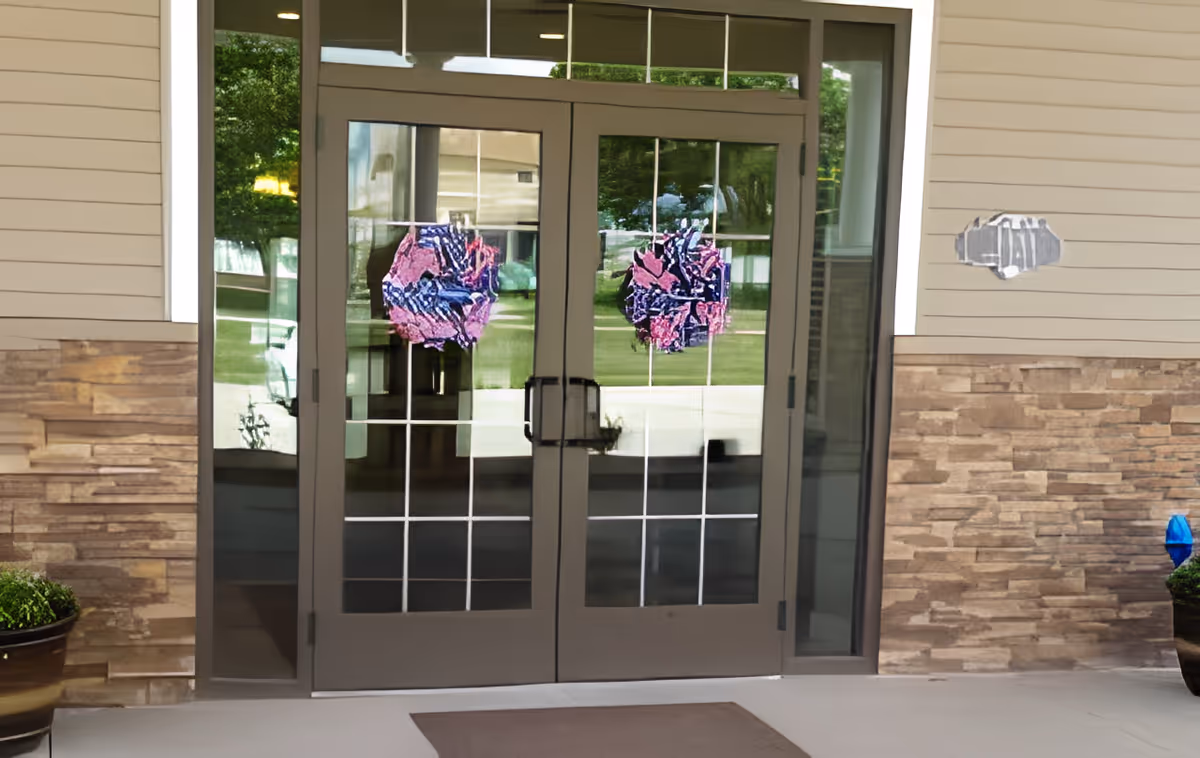 Glass double doors with grid patterns and decorative wreaths, set in a building entrance with stone and beige siding walls, and potted plants on either side.