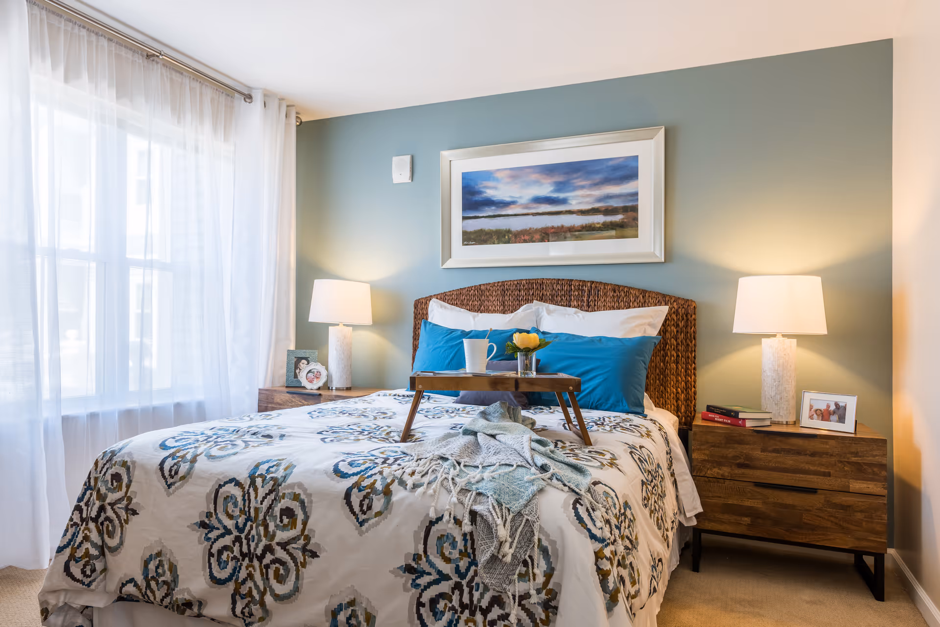 Well-lit bedroom with a patterned bedspread, blue pillows, a breakfast tray, bedside tables with lamps, and a framed landscape above the wicker headboard.