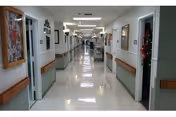 A long, clean hallway inside a nursing and rehab center with handrails on both sides, doors leading to rooms, and framed pictures on the walls. The floor is shiny and reflective, and the corridor is well-lit with ceiling lights.