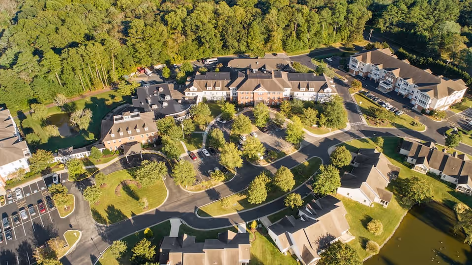 Aerial view of Lakeside At Mallard Landing senior living facility showing multiple buildings surrounded by trees, parking lots, roads, and small ponds.