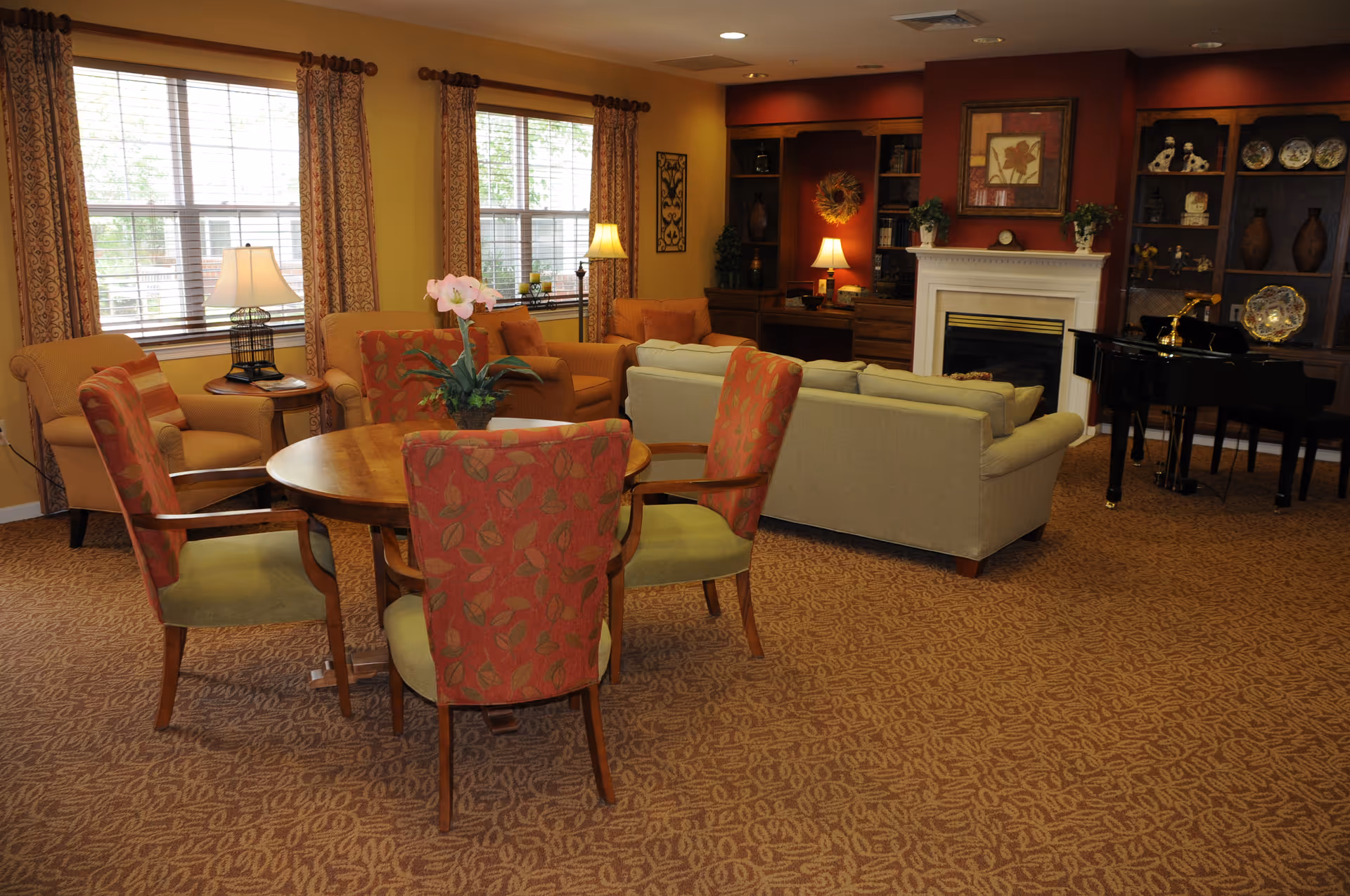 Cozy communal living room with a round table and upholstered chairs, sofas facing a fireplace, built-in bookshelves, and a piano.