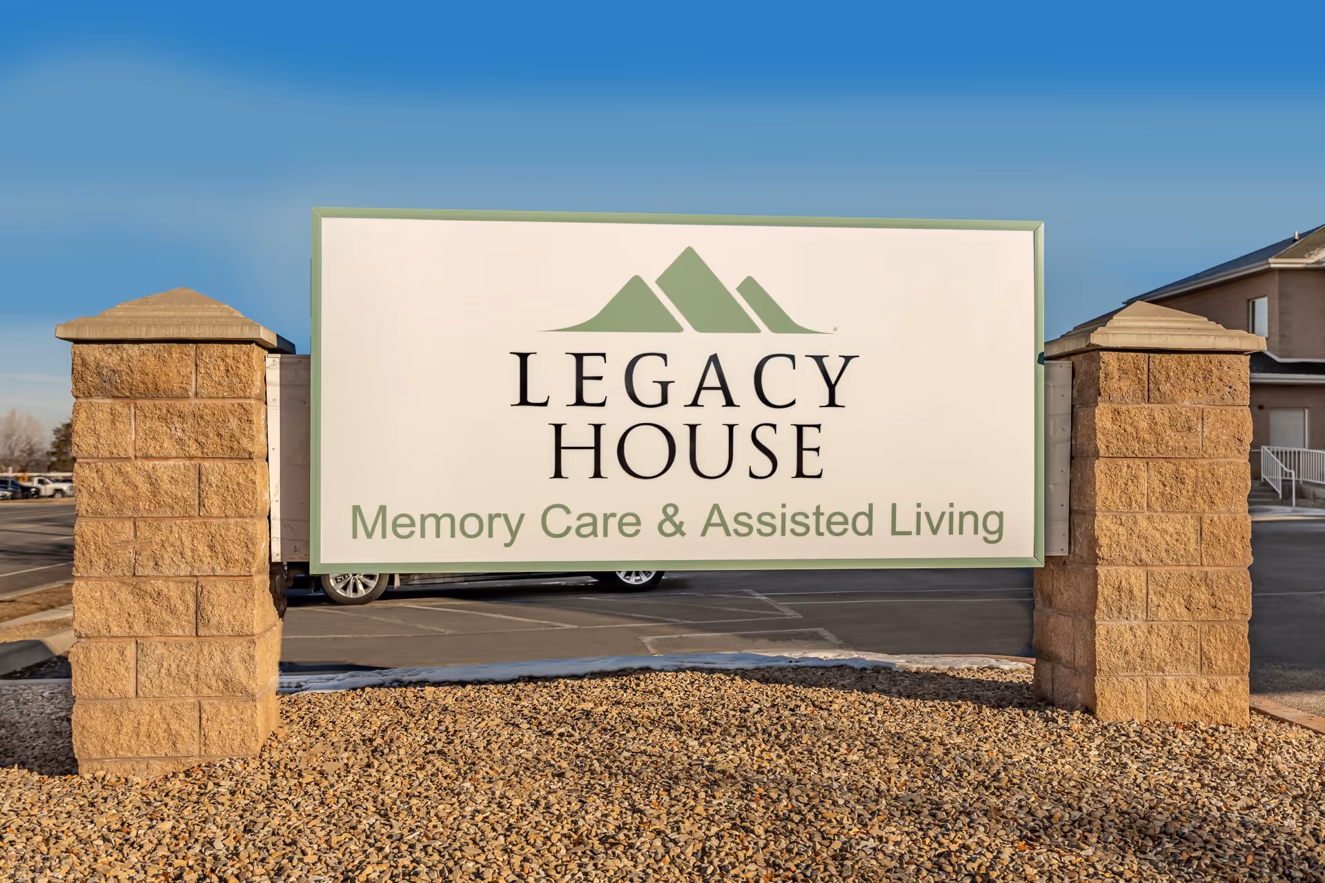 Outdoor sign for Legacy House Memory Care & Assisted Living mounted between two stone pillars with a parking lot and building in the background under a clear blue sky.