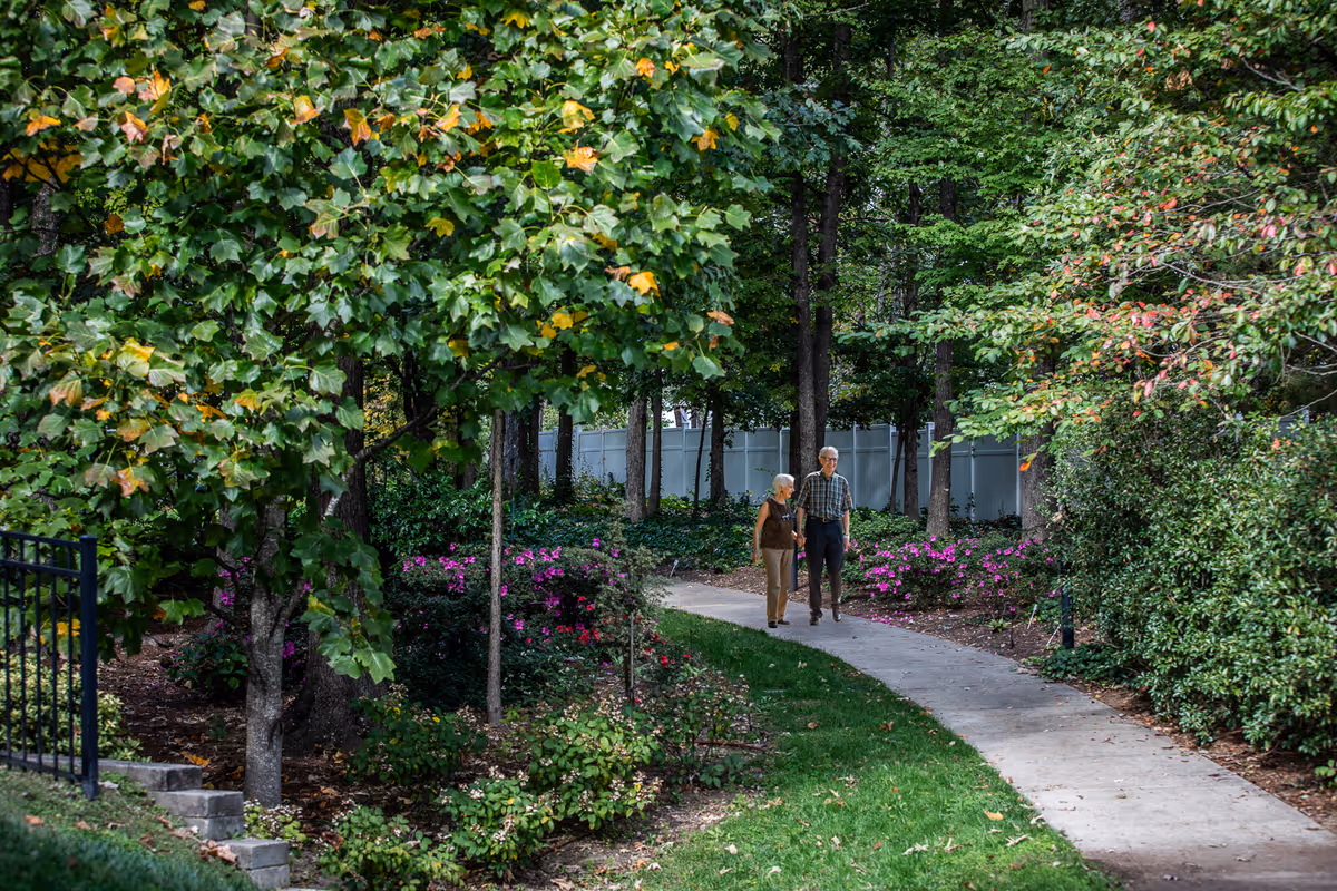 An elderly couple walking together on a curved concrete pathway surrounded by lush green trees and flowering bushes in a garden area.