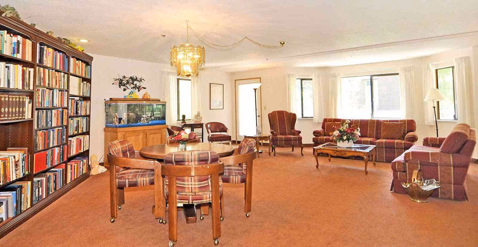 A spacious living room with a large bookshelf filled with books on the left, a fish tank on a wooden cabinet, a round wooden table with four plaid cushioned chairs, and a seating area with a striped sofa, armchairs, and a coffee table with a flower arrangement. The room has large windows with white curtains and warm lighting.