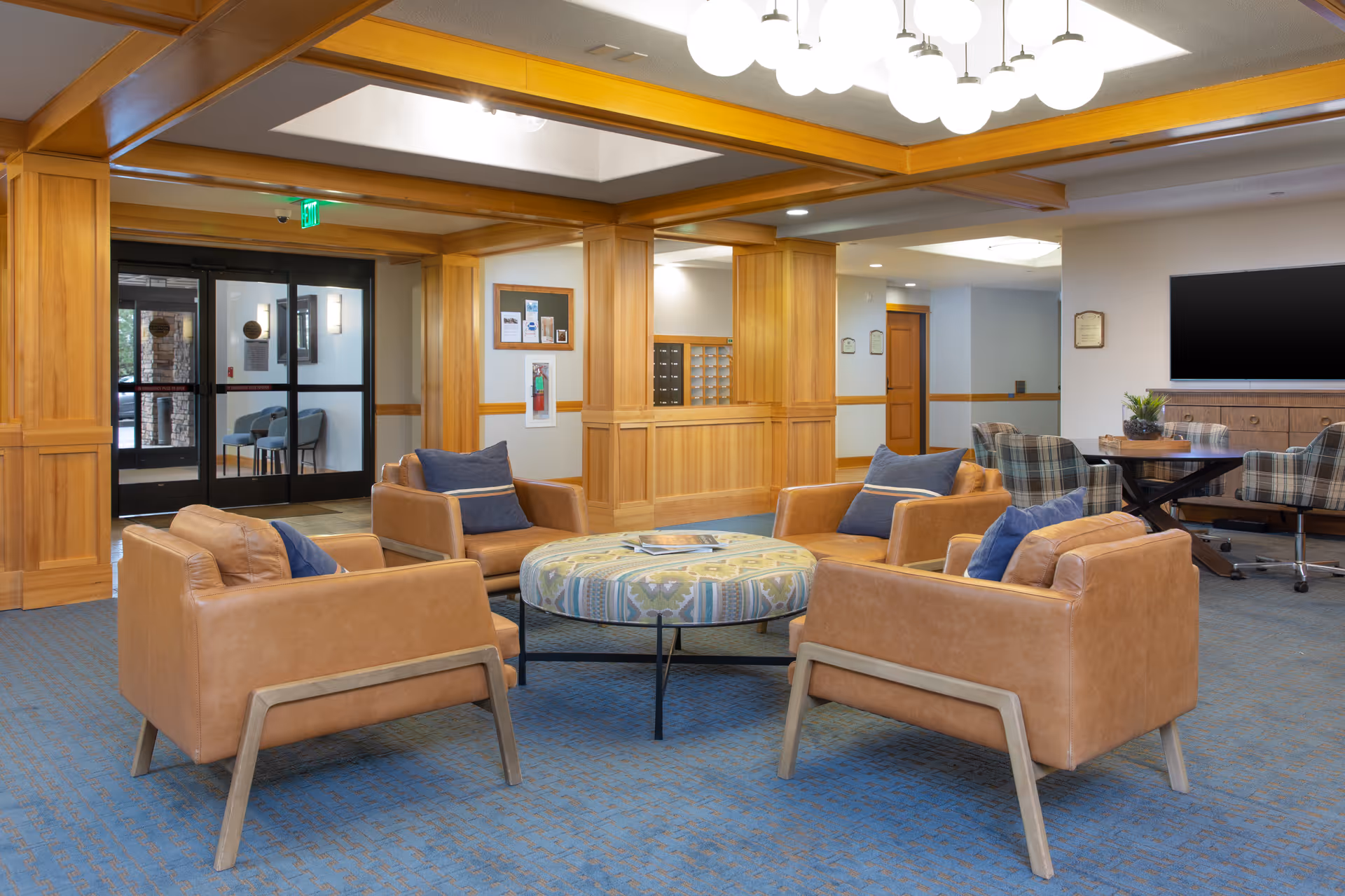 A spacious and well-lit common area with four tan leather armchairs arranged around a round patterned ottoman. The room features wooden paneling, a blue carpet, a large flat-screen TV mounted on the wall, and a table with plaid upholstered chairs in the background. Glass double doors lead to another area, and there is a bulletin board and mailboxes on the wall.