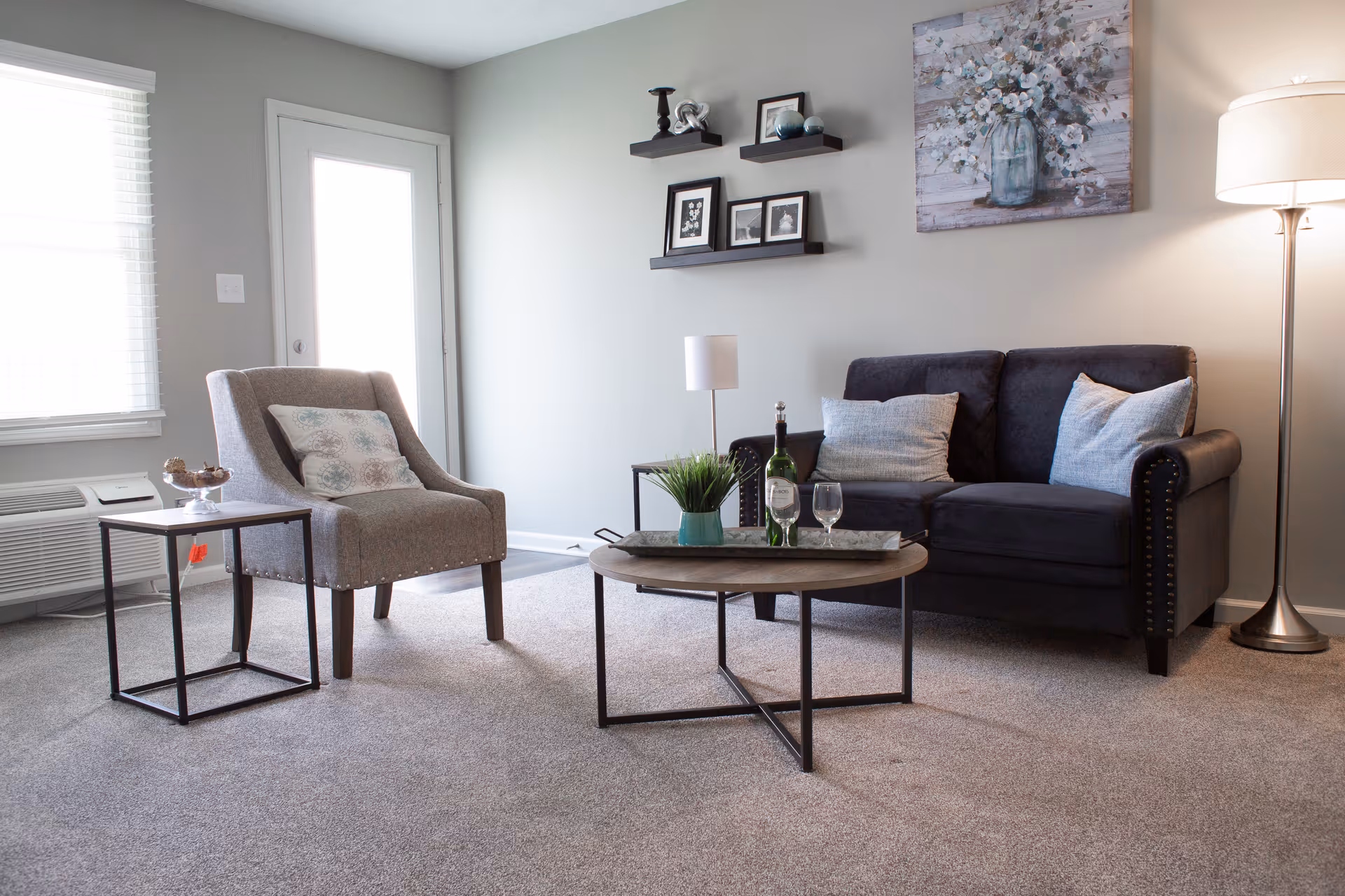 A cozy living room with a gray armchair and a dark gray loveseat with light-colored pillows. There is a round wooden coffee table with a plant, a bottle of wine, and two wine glasses. A floor lamp stands in the corner, and a painting of white flowers in a vase hangs on the wall above the loveseat. Three small shelves with framed pictures and decorative items are mounted on the wall. A window with blinds and a door are visible in the background.