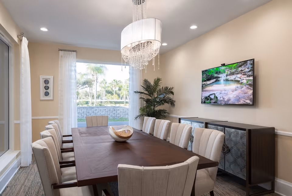 A bright dining room with a long wooden table surrounded by upholstered chairs, a crystal chandelier, a wall-mounted TV and sideboard, and a large window showing palm trees outside.