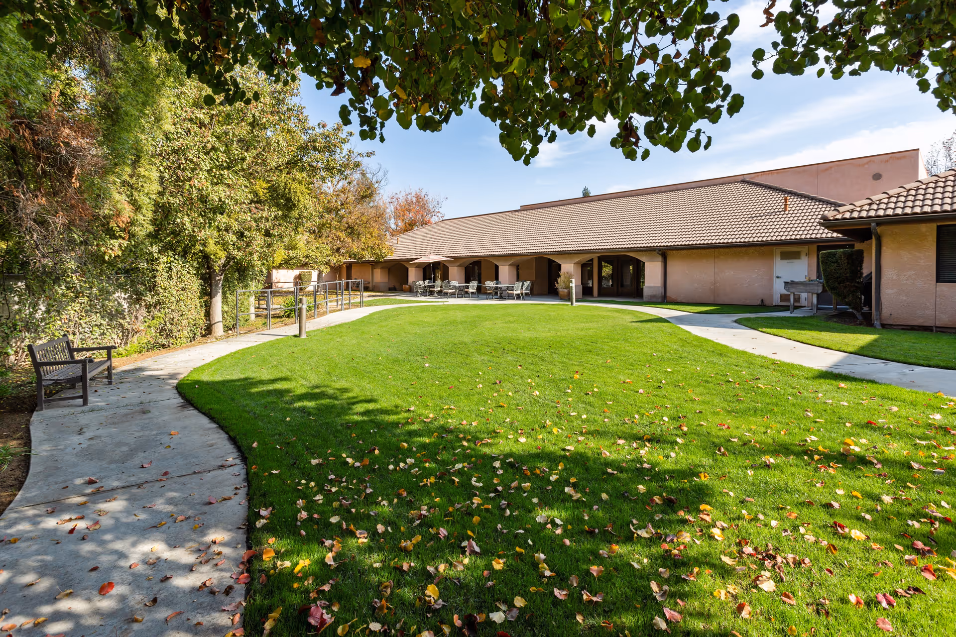 A well-maintained outdoor garden area at Summerfield Memory Care Of Fresno featuring a green lawn with scattered autumn leaves, a curved concrete walkway, benches along the path, and a building with a tiled roof and covered patio seating in the background under a partly cloudy sky.
