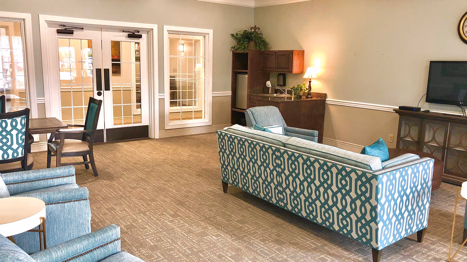 A cozy lobby seating area featuring patterned chairs and a small table, with glass doors leading to another room and a kitchenette in the background.