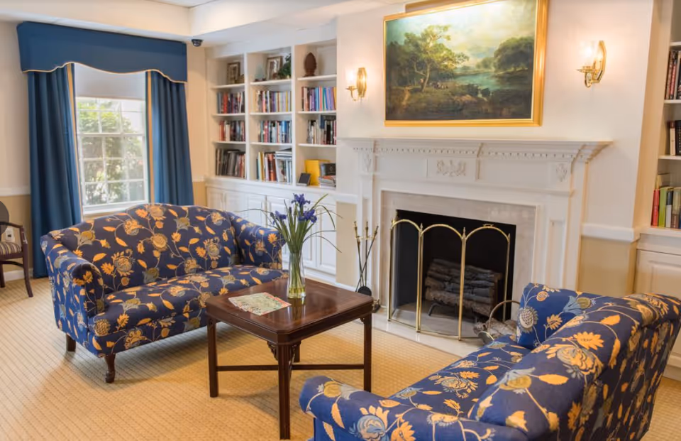 A cozy living room with two blue floral-patterned sofas facing each other across a wooden coffee table with a vase of purple flowers. The room features a white fireplace with a brass screen, a large framed landscape painting above it, built-in white bookshelves filled with books, and a window with blue curtains.