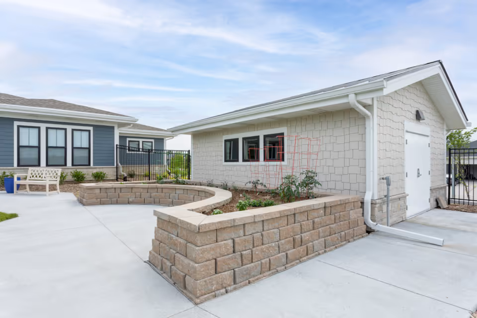Outdoor courtyard area of a senior living facility with a raised garden bed made of stone bricks, a white bench, and two buildings with windows and light-colored siding under a partly cloudy sky.