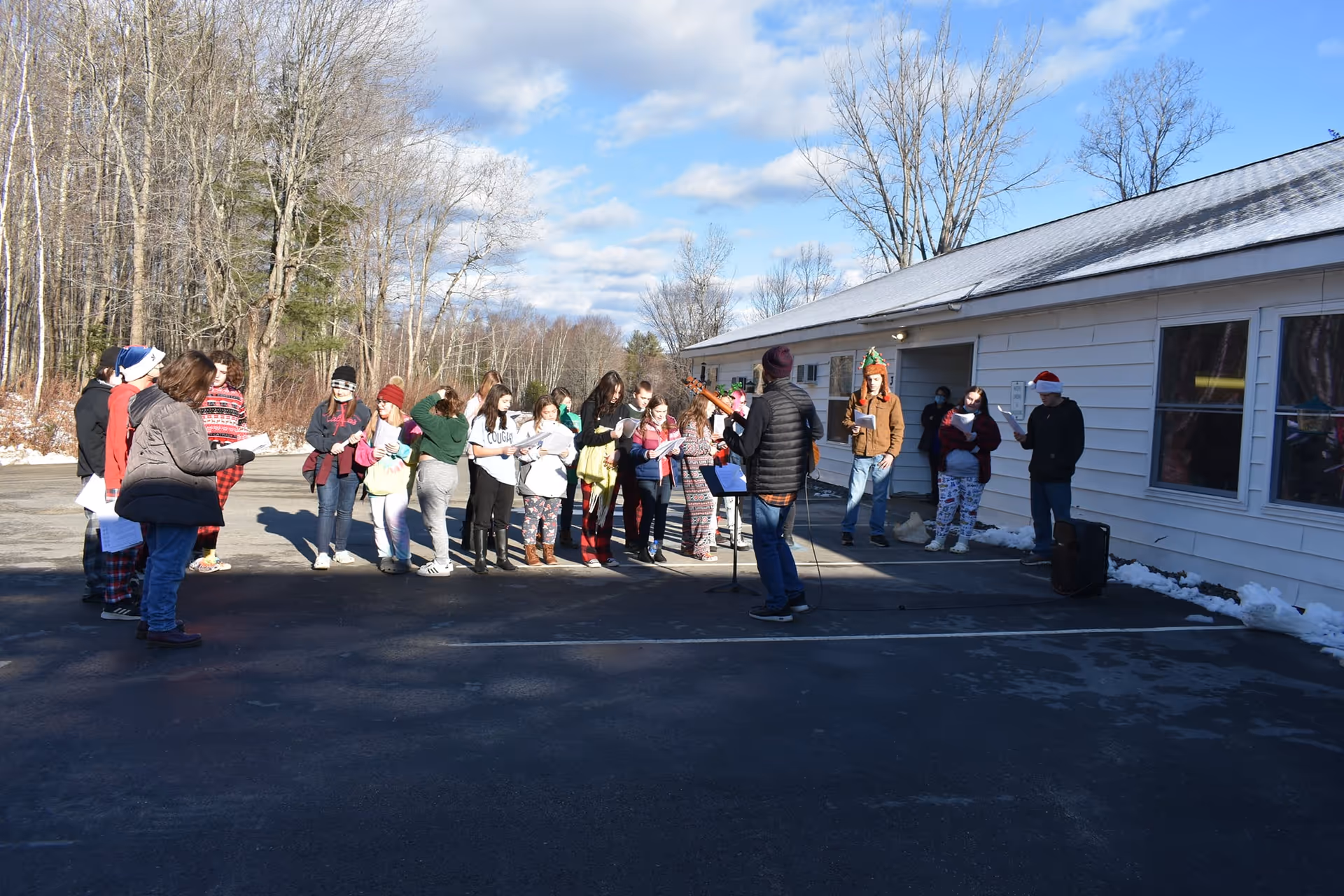 A group of people standing outside in a parking lot next to a white building. Most are holding papers and appear to be singing or rehearsing, with one person playing a guitar. It is a sunny day with some snow on the ground and leafless trees in the background.