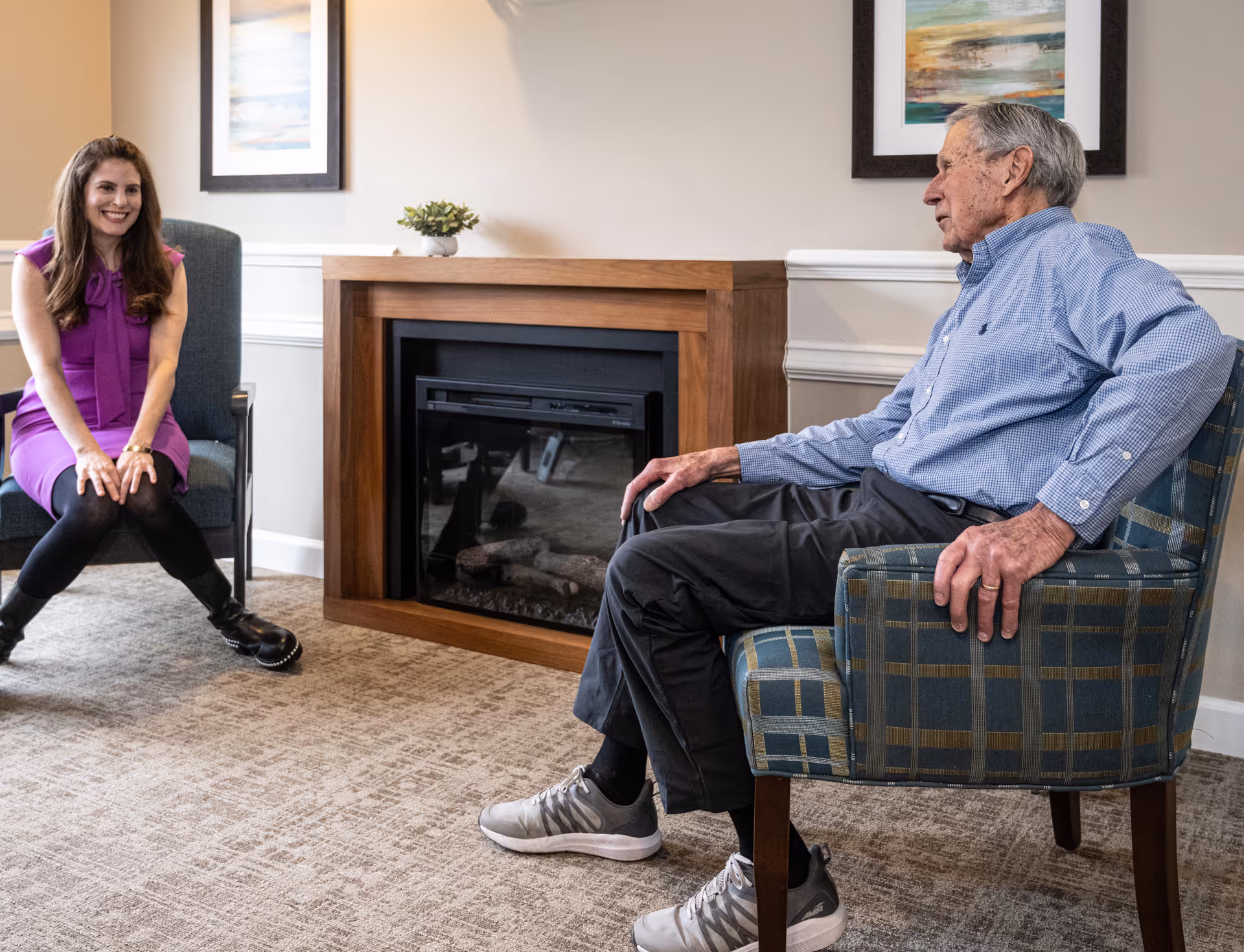 An elderly man and a young woman sitting in armchairs facing each other in a cozy room with a fireplace between them. The man is wearing a blue checkered shirt and dark pants, while the woman is dressed in a purple dress with black tights and boots. The room has beige walls, carpeted floor, and framed artwork above the fireplace.