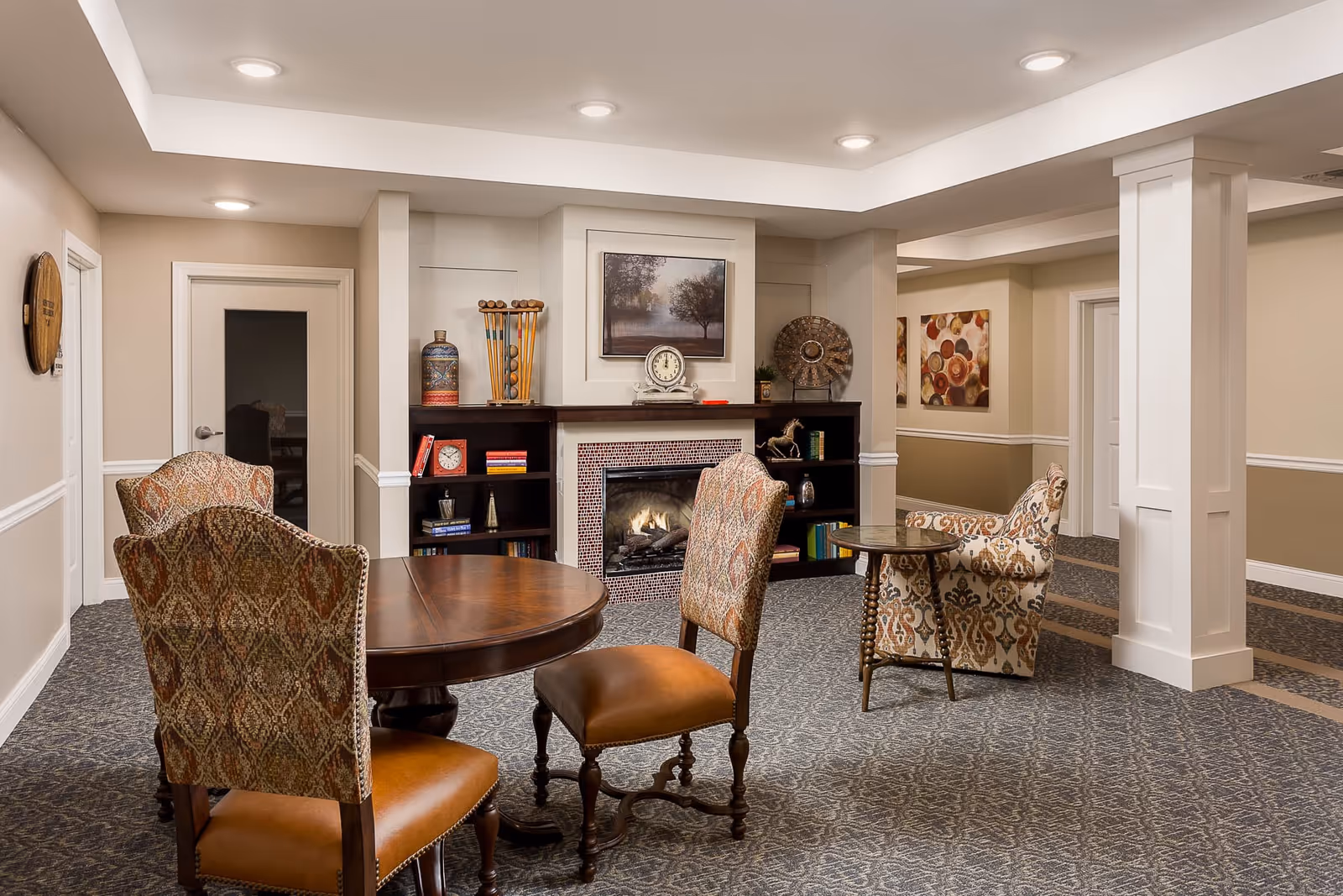 A cozy senior living common area with patterned upholstered chairs around a round wooden table and a small side table. The room features a lit fireplace with decorative items on the mantel and built-in shelves on either side. The walls are beige with white trim, and there are framed artworks hanging on the walls. The carpet has a subtle pattern, and recessed ceiling lights illuminate the space.