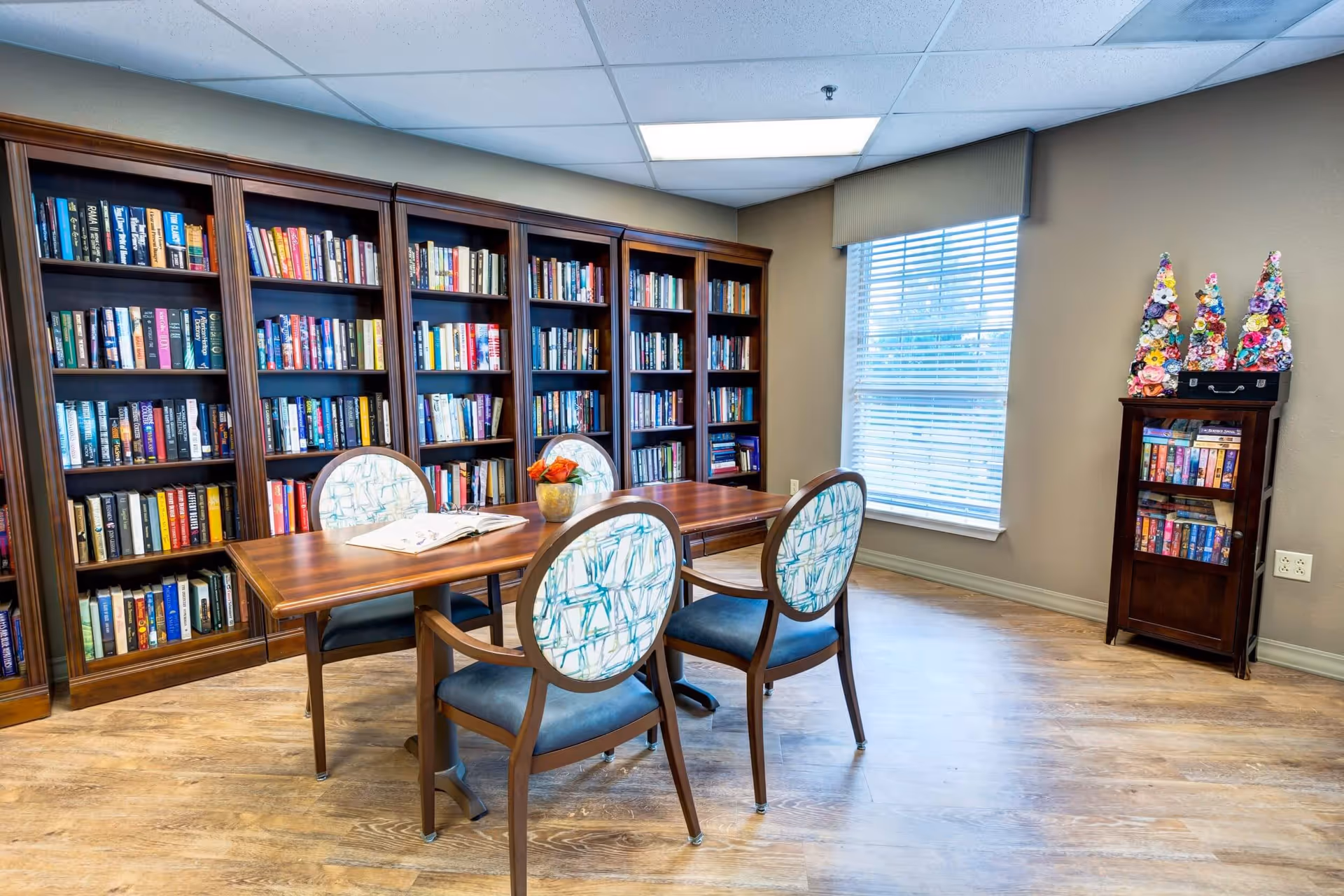 A cozy library room with wooden bookshelves filled with books along the wall. In the center, there is a wooden table with four chairs featuring patterned backs and blue seats. A small vase with orange flowers is placed on the table. A window with blinds allows natural light into the room, and a small wooden cabinet with decorative floral cone-shaped ornaments is positioned near the window.