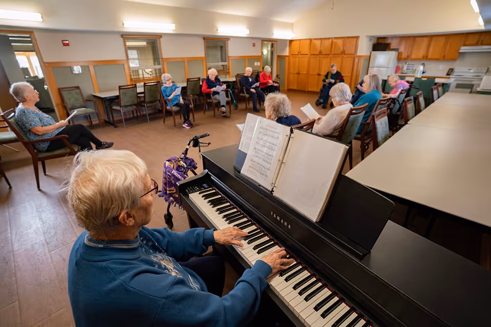 A group of elderly people seated in a semi-circle in a senior living facility room, with one person playing a Yamaha piano in the foreground. The room has wooden floors, tables, chairs, and kitchen cabinets in the background.