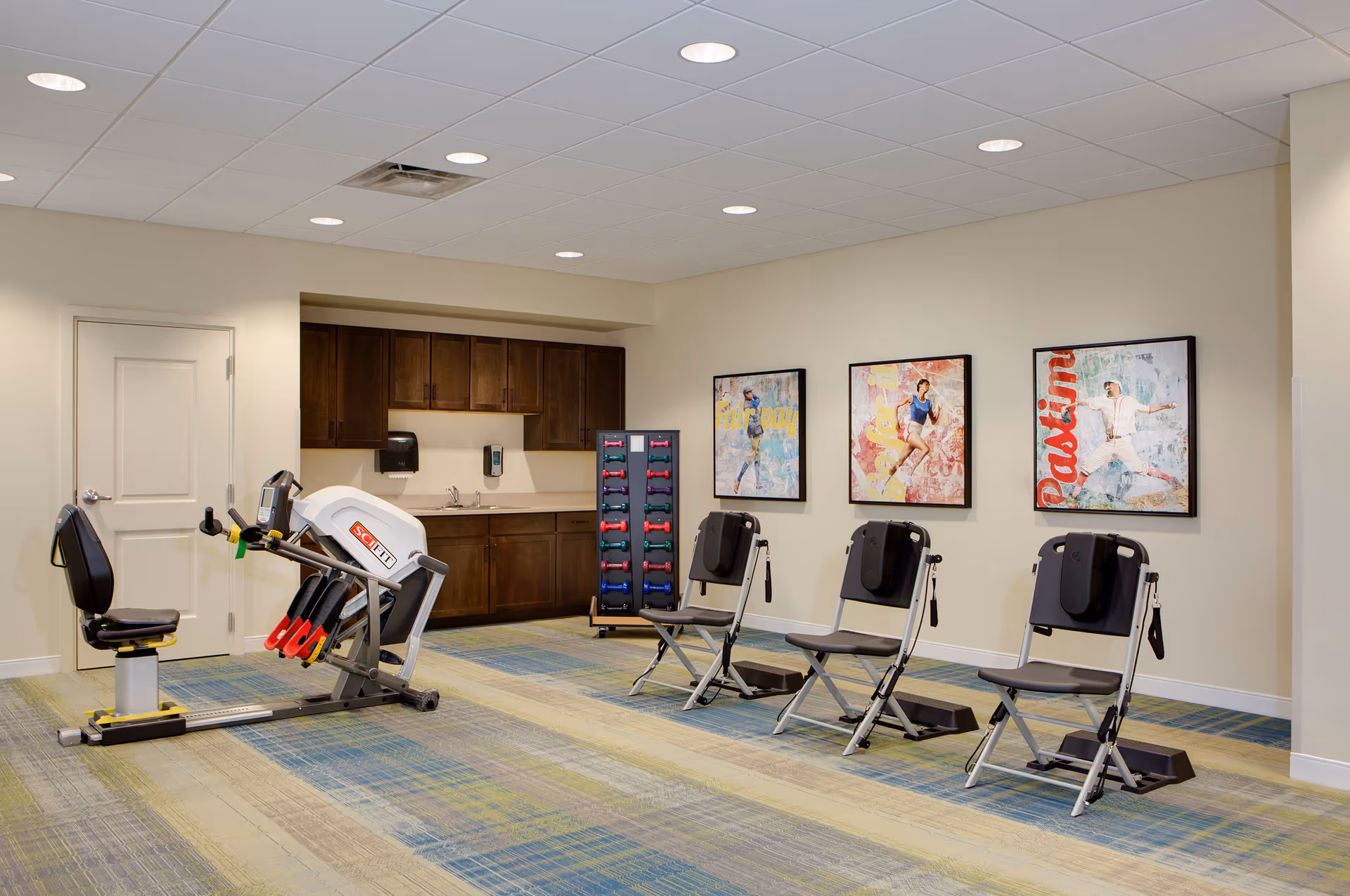 A fitness room in Prairie House featuring exercise equipment including a recumbent exercise machine, three black exercise chairs with footrests, a rack of colorful dumbbells, and a kitchenette area with wooden cabinets and a sink. The walls are decorated with three framed sports-themed artworks.
