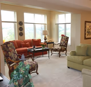 A bright living room with large windows letting in natural light. The room features a red sofa, two floral-patterned armchairs, a green sofa, a coffee table with flowers and a wine bottle, a side table with a lamp, and decorative wall art. There is a potted plant in the foreground.