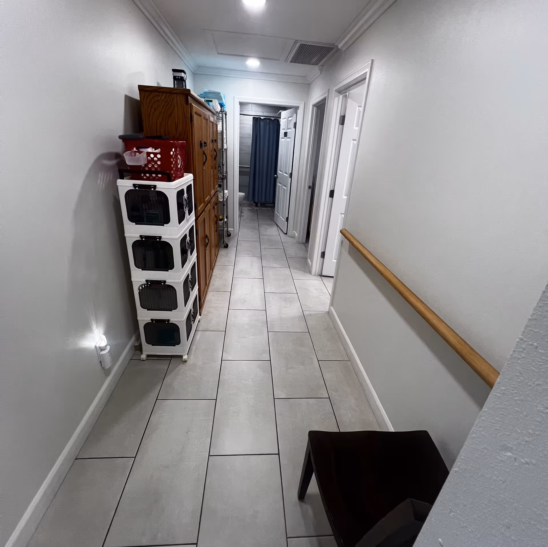 A narrow hallway with light gray tiled floor and white walls. On the left side, there is a tall wooden cabinet and a white storage unit with multiple compartments. On the right side, there is a wooden handrail attached to the wall and a dark-colored chair near the foreground. At the end of the hallway, there are three doors, one of which is open revealing a bathroom with a toilet and a blue shower curtain.
