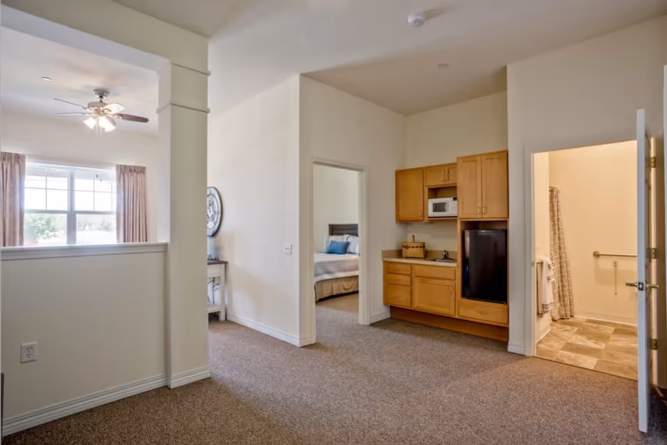 Open senior living apartment interior with a small kitchenette, a doorway to a bedroom, and a bathroom visible to the right.