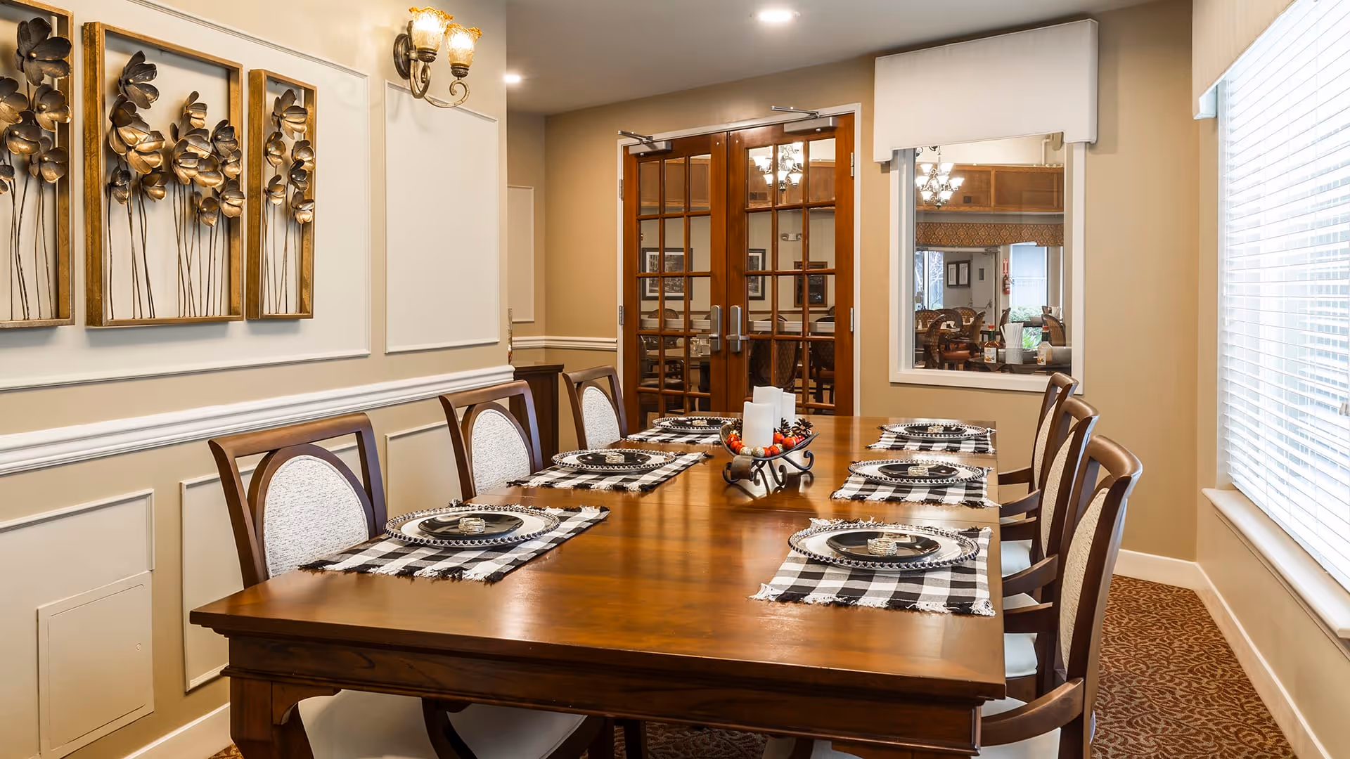 A well-lit dining room with a long wooden table set for eight people. Each place setting has a black and white checkered placemat, a decorative plate, and a small candle. The room features beige walls with white trim, decorative metal wall art of flowers, a window with blinds on the right, and double glass doors at the far end.