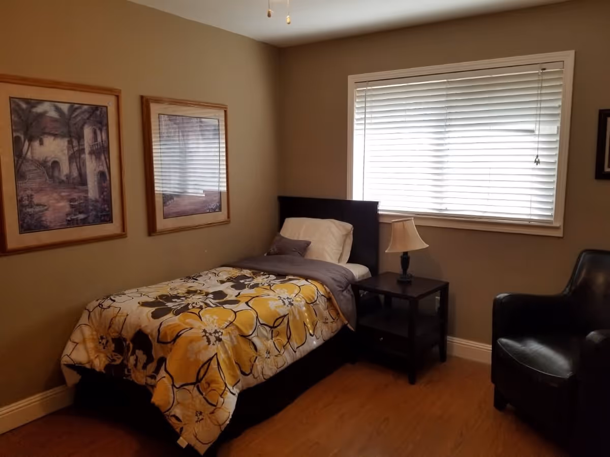 A small bedroom with a single bed covered in a yellow and gray floral comforter, a white pillow, and a gray blanket. Next to the bed is a black nightstand with a lamp on it. There is a black armchair to the right of the nightstand. The room has beige walls with two framed pictures hanging on the left wall and a window with white blinds on the right wall. The floor is wooden.