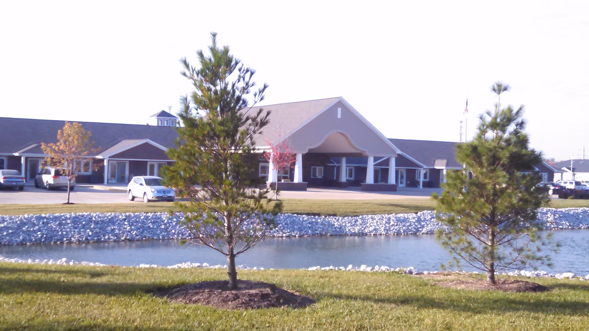 Exterior view of Villas of Holly Brook Assisted Living facility in Washington, IL, showing a large building with a covered entrance, a parking lot with several cars, a small pond bordered by white rocks, and two pine trees in the foreground on a grassy area.