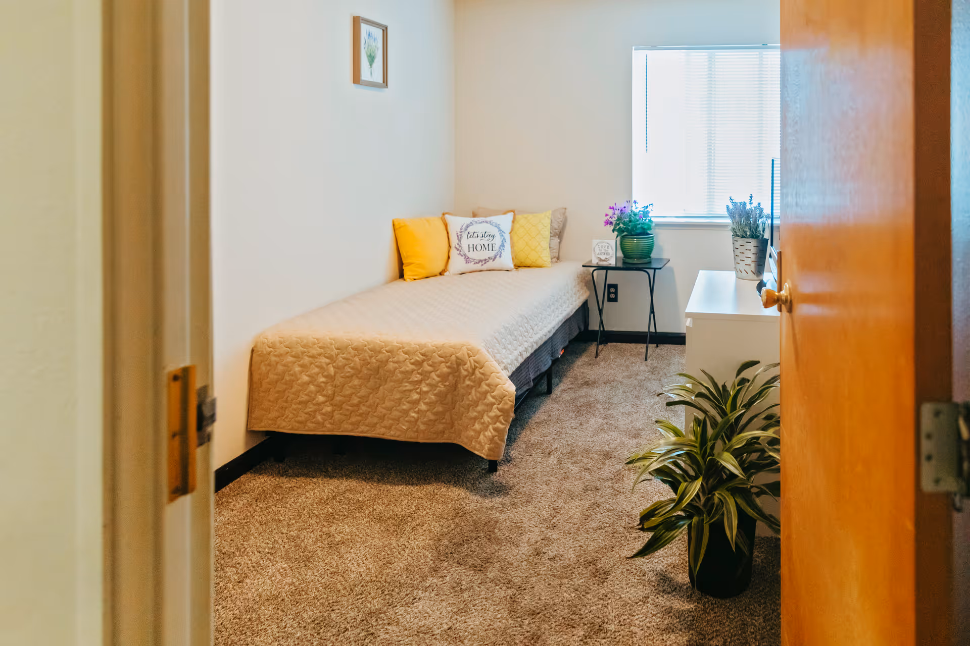 A small, cozy bedroom with a single bed covered in a beige quilt and decorated with yellow and patterned pillows. There is a small black side table next to the bed with a green potted plant and a decorative item. A window with blinds lets in natural light. The room has beige carpet flooring, a wooden door partially open, and a green leafy plant on the floor near a white dresser.
