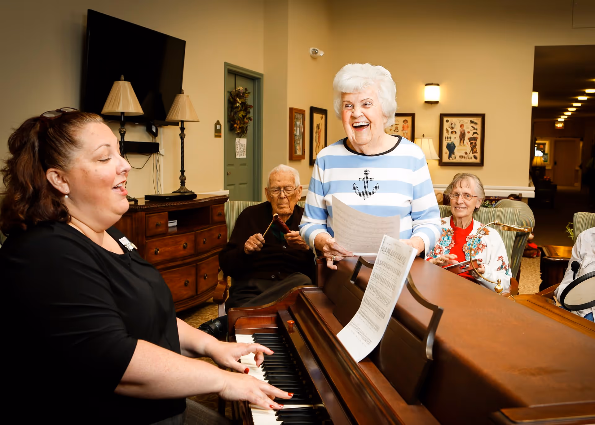 A group of elderly people enjoying a music session in a common area. One woman is playing the piano while another woman stands beside her holding sheet music and smiling. Two other elderly individuals are seated nearby, one playing a percussion instrument and the other holding a tambourine. The room has warm lighting, framed pictures on the walls, and a television mounted above a wooden cabinet.