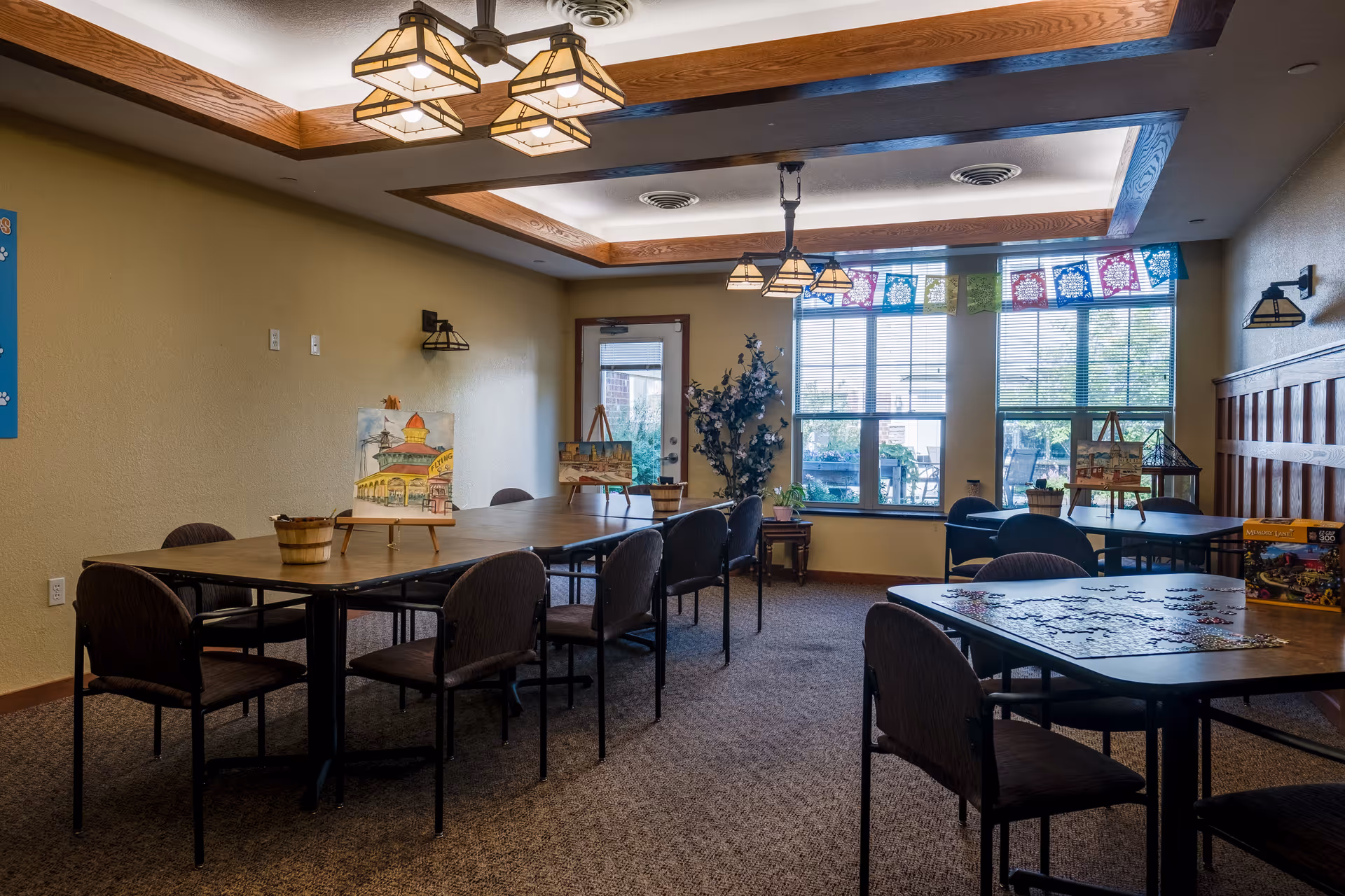 A well-lit activity room with multiple tables and chairs arranged for group activities. The tables have art supplies and paintings on easels, as well as a partially completed jigsaw puzzle. Large windows with colorful paper decorations allow natural light to fill the room. The ceiling features wooden beams and hanging light fixtures.