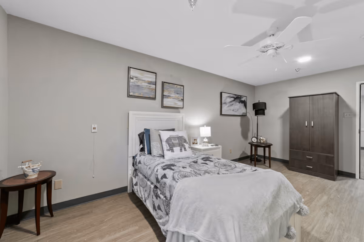 A neatly arranged bedroom with a single bed featuring a patterned bedspread and an elephant-themed pillow. The room has light gray walls with three framed artworks above the bed. There is a white nightstand with a lamp and picture frames, a dark wooden wardrobe, a small round table with a black lamp, and a ceiling fan. The floor is light wood, and the room has a calm, neutral color palette.