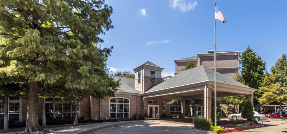 The covered entrance of a brick senior living building with a porte-cochere, flagpole, and surrounding trees.