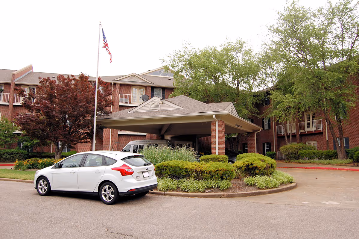 Exterior view of Kennington Pointe senior living facility showing a brick building with balconies, a covered entrance with a peaked roof, a white car parked in front, an American flag on a flagpole, and surrounding trees and shrubs.