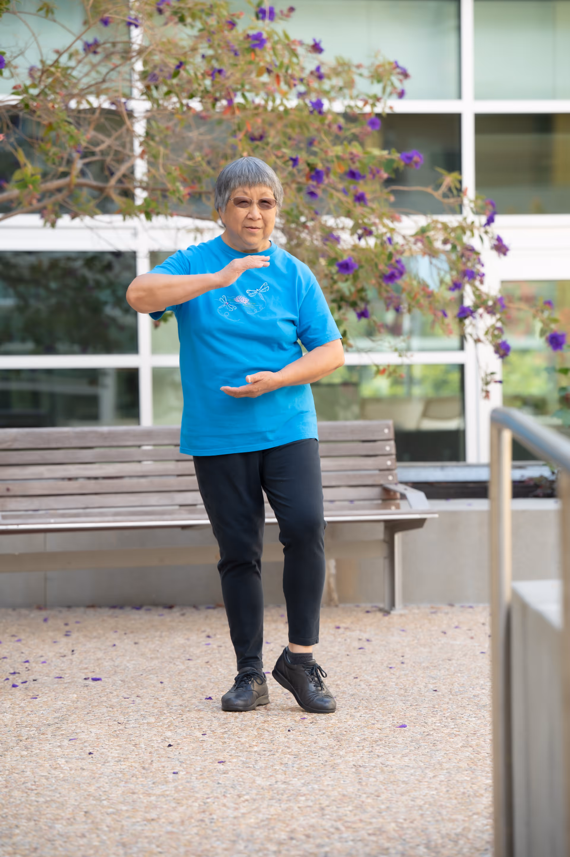 An elderly woman wearing a blue t-shirt and black pants is practicing Tai Chi outdoors in front of a bench and a building with large windows. Purple flowers and green foliage are visible in the background.