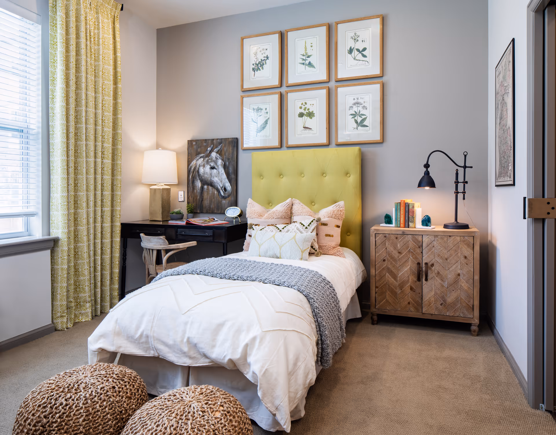 A cozy bedroom with a single bed featuring a yellow tufted headboard, white bedding, and multiple decorative pillows. There is a wooden nightstand with a black lamp and books on it to the right of the bed. On the left side, there is a black desk with a lamp, a chair, and a horse painting hanging above it. Six framed botanical prints are arranged in two rows above the headboard. The room has beige carpet, yellow patterned curtains, and a window letting in natural light. Two woven poufs are placed at the foot of the bed.