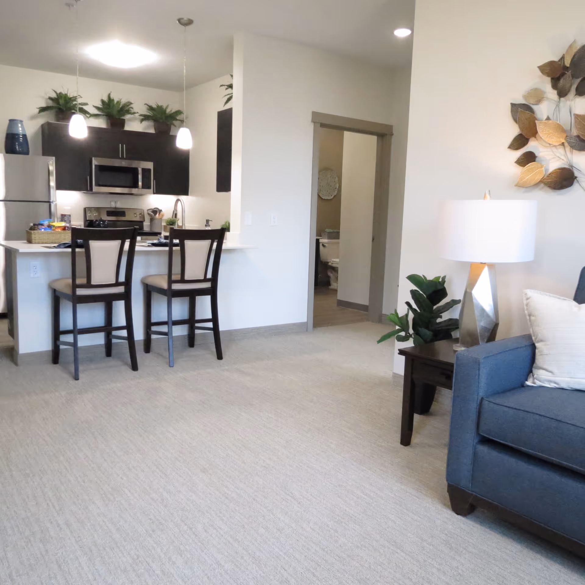 Interior view of a senior living facility apartment showing a small kitchen area with two chairs at a counter, stainless steel refrigerator and microwave, dark cabinets, and potted plants on top. To the right is a living area with a blue armchair, side table with a lamp, and decorative wall art. A doorway leads to a bathroom with a visible toilet.