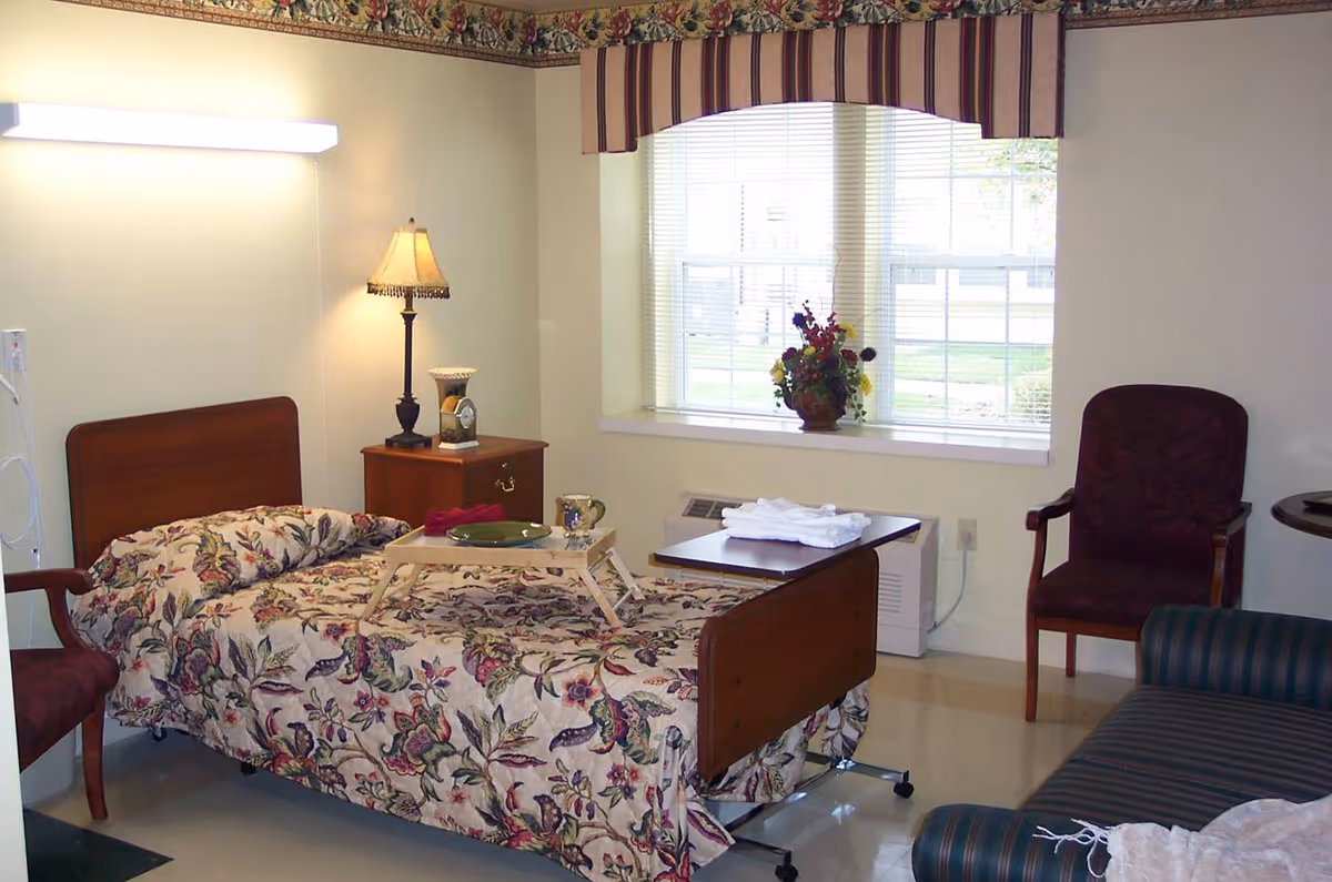 A senior living facility bedroom with a single bed covered in floral patterned bedding. There is a wooden nightstand with a lamp and a clock, a tray with a plate and cup on the bed, a window with blinds and a striped valance, a floral arrangement on the windowsill, two chairs, and a striped couch with a blanket.