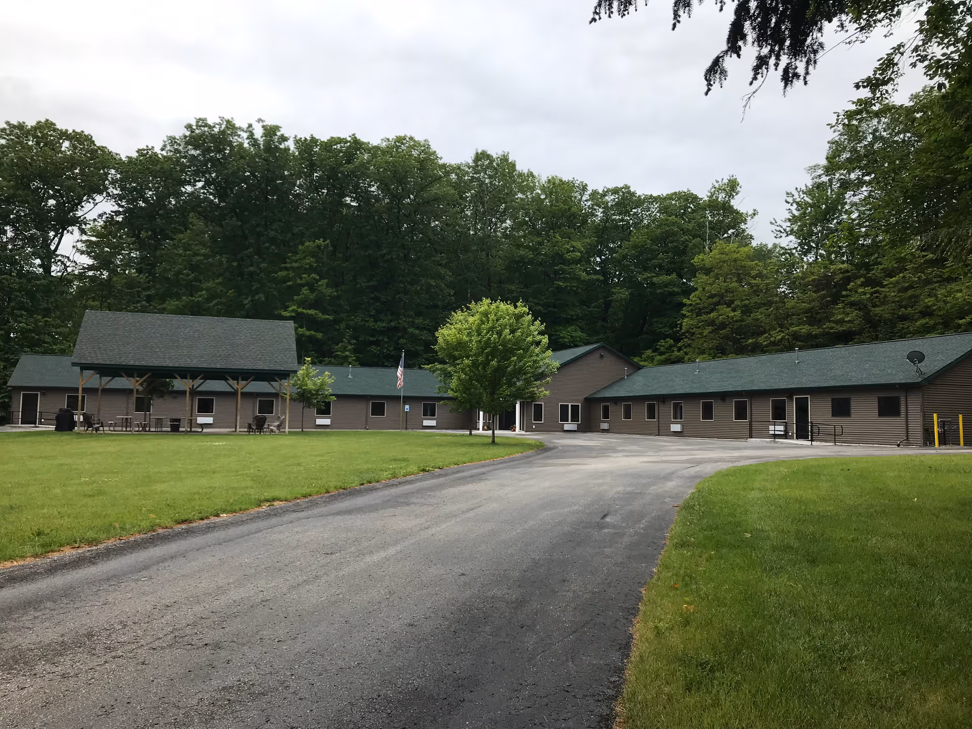 A single-story assisted living facility building with a green roof surrounded by a large grassy area and trees in the background. There is a paved driveway leading up to the building, and a covered outdoor seating area with tables and chairs is visible on the left side.