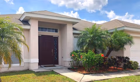 Front exterior view of a single-story residential building with a dark brown front door, beige walls, a sloped roof, and landscaped greenery including palm trees and flowering bushes under a partly cloudy sky.