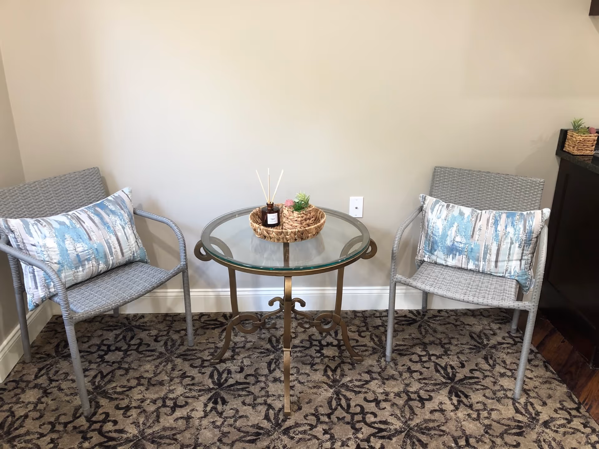 Two gray wicker chairs with blue and white patterned cushions are placed on either side of a small round glass-top table with a decorative metal base. On the table, there is a woven tray holding a small plant and a reed diffuser. The setting is indoors with a beige wall and patterned carpet.