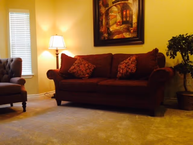 A cozy living room with a dark red sofa adorned with patterned cushions, a beige armchair, a floor lamp emitting warm light, a framed painting on the wall, and a potted plant in the corner near a window with blinds.