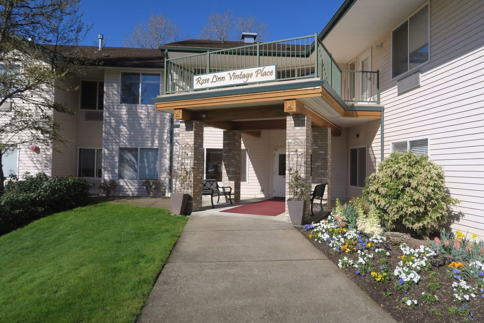 Exterior view of Rose Linn Vintage Place, showing the entrance with a covered porch supported by brick columns, a red carpet leading to the door, benches on either side, and a garden with colorful flowers and green grass on the right side.