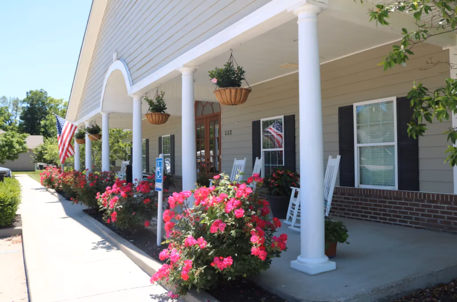 Front porch of a senior living facility with white columns, hanging flower baskets, vibrant pink flowers along the walkway, white rocking chairs, and an American flag displayed near the entrance.