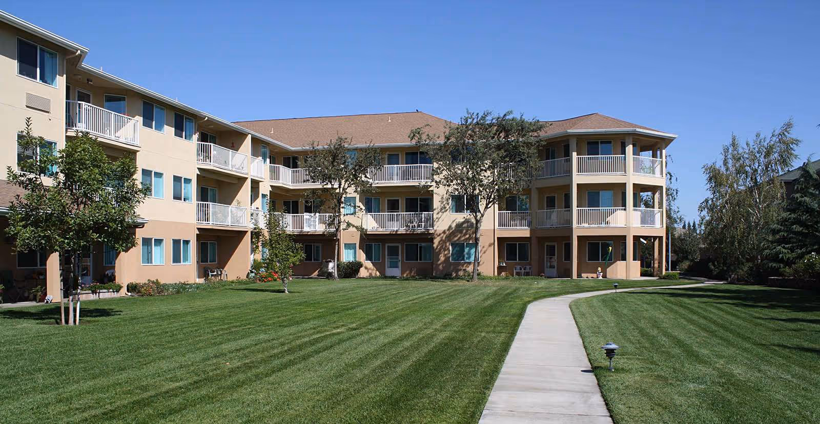 Exterior view of a three-story senior living facility building with balconies, surrounded by a well-maintained lawn, trees, and a concrete walkway leading to the entrance under a clear blue sky.
