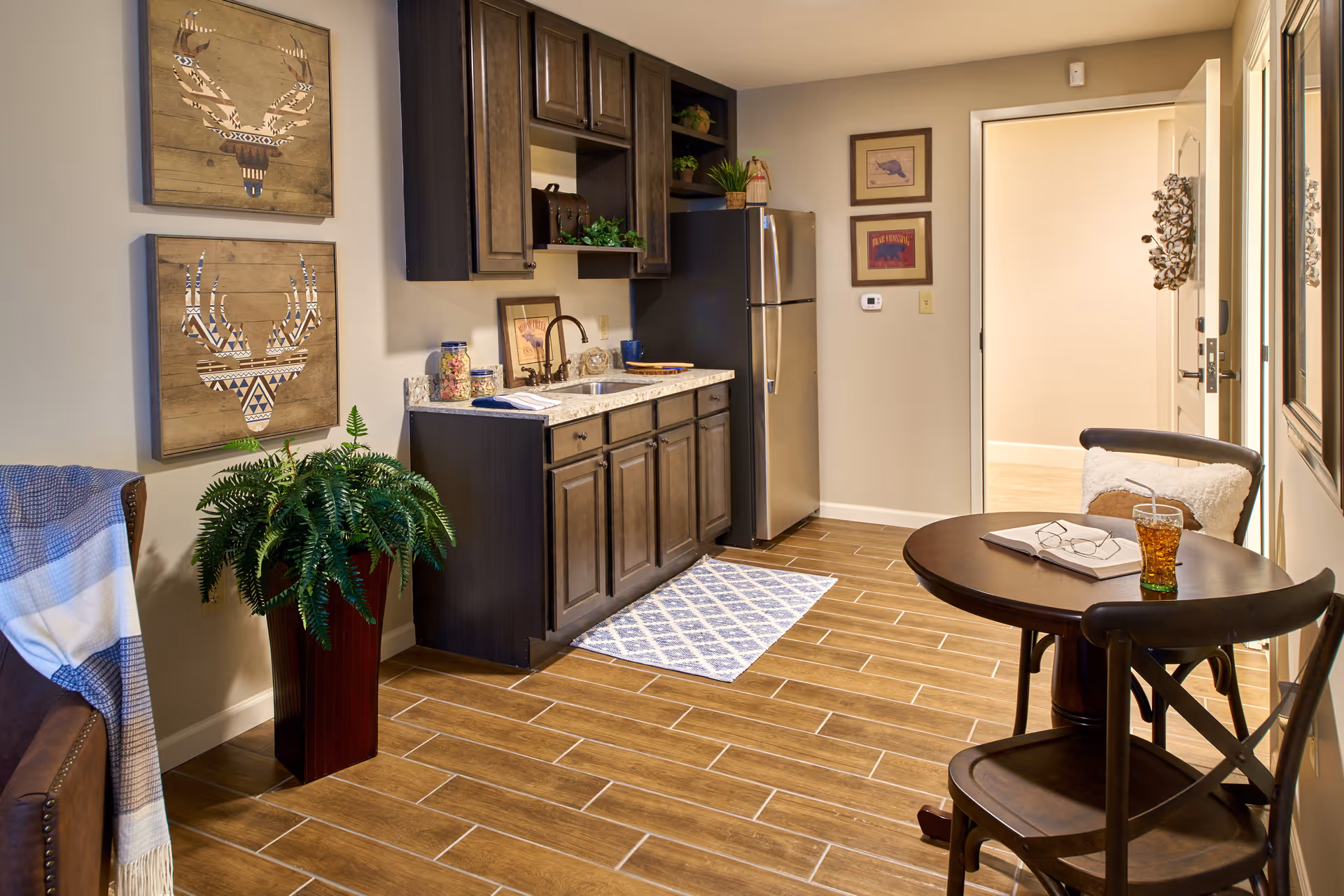 A cozy kitchen area with dark wooden cabinets, a stainless steel refrigerator, a small sink, and a granite countertop. There is a round wooden table with two chairs, one with a cushion and a glass of iced tea on the table. The floor has wood-like tiles, and the walls are decorated with framed artwork and two wooden deer head wall hangings. A green potted plant is placed near a brown armchair with a blue and white blanket draped over it.