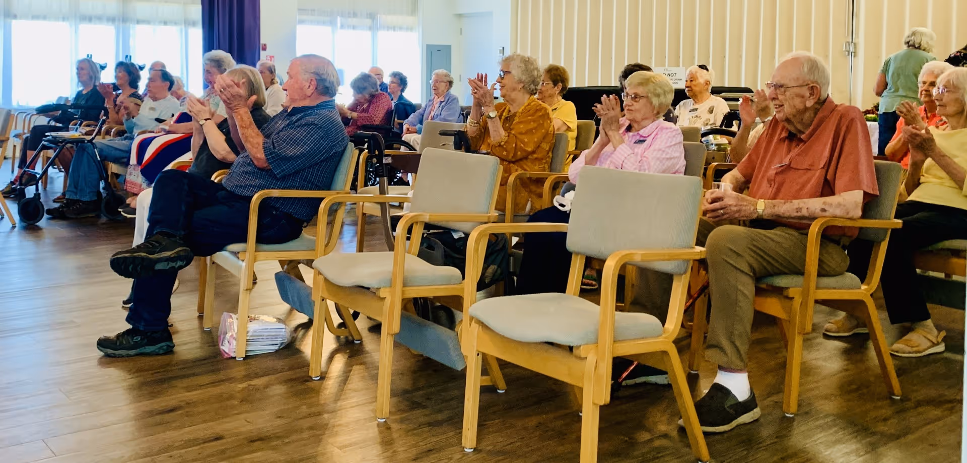 A group of elderly people sitting in chairs arranged in rows inside a well-lit room, clapping and engaging in an event or activity. The room has wooden flooring and large windows with curtains allowing natural light to enter.