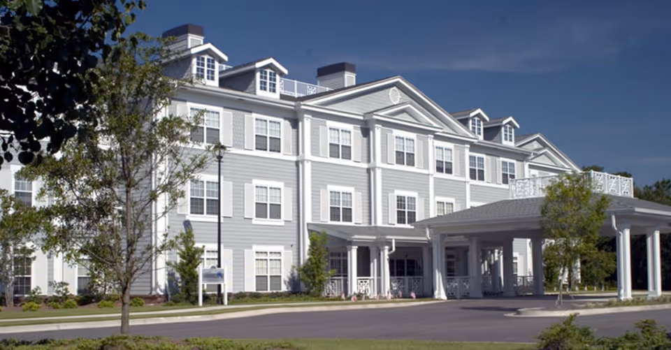 Exterior view of a large, three-story senior living facility building with light gray siding and white trim, multiple windows, and a covered entrance driveway. The building is surrounded by trees and landscaping under a clear blue sky.